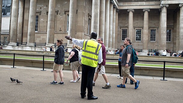A security officer guides a group of people towards the left hand side of the photograph. In the background, people queing in front of a museum can be seen.