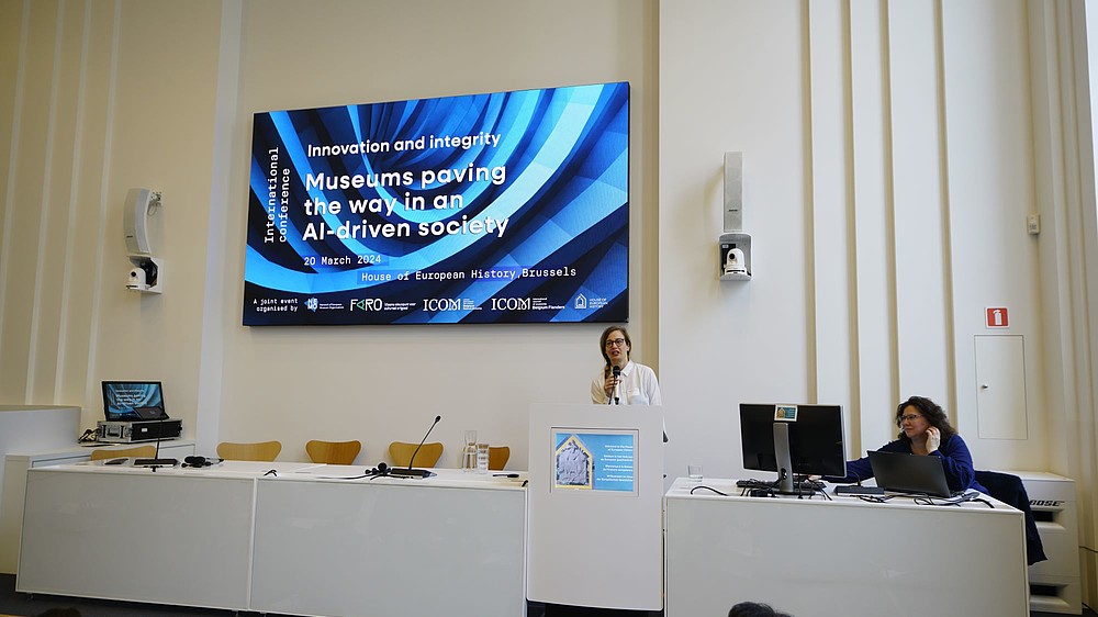 A person is standing behind a podium talking into a handheld microphone. Behind her there is a projected image with the name of the conference "Innovation and Integrity: Museums paving the way in an AI-driven society".  © Image: Peter Van der Plaetsen